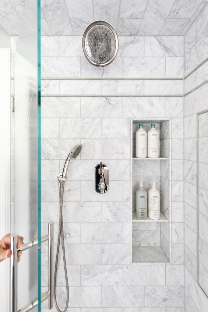 Marble-tiled shower with a rain showerhead, handheld shower, and niche holding bottles. A hand opens the glass door. Minimalist and clean design.