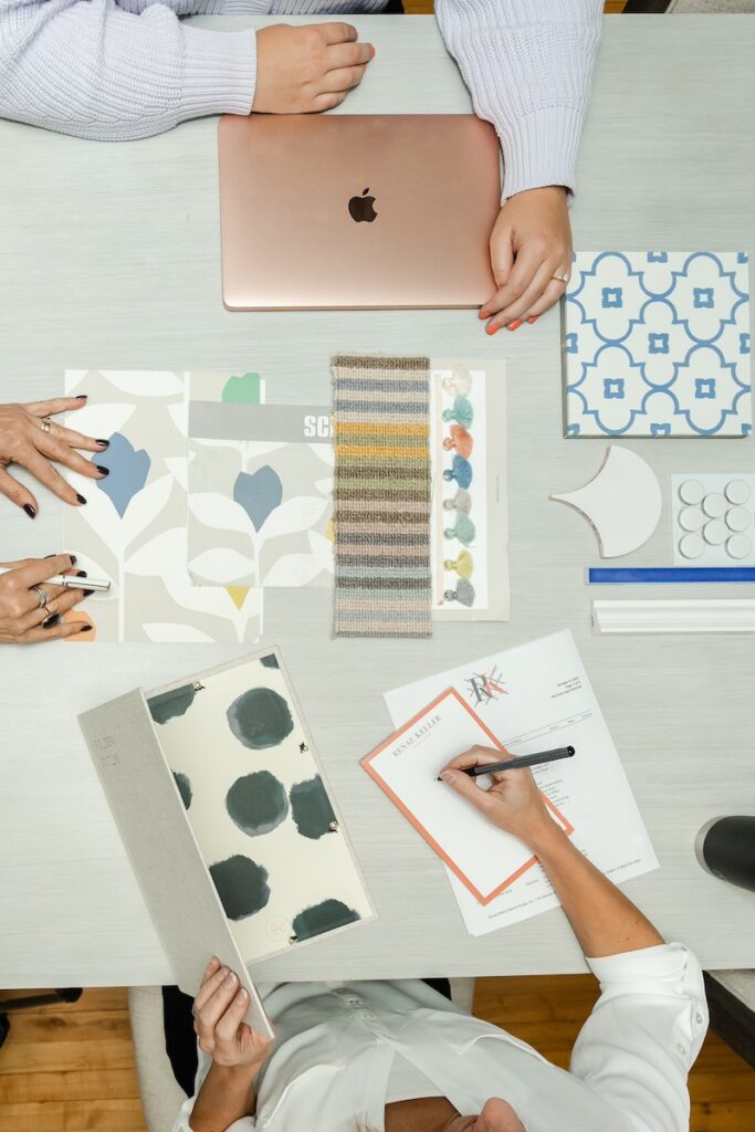 Three people view patterns and a laptop at a table. Papers and fabric swatches are spread out. Neutral tones with a patterned blue tile.