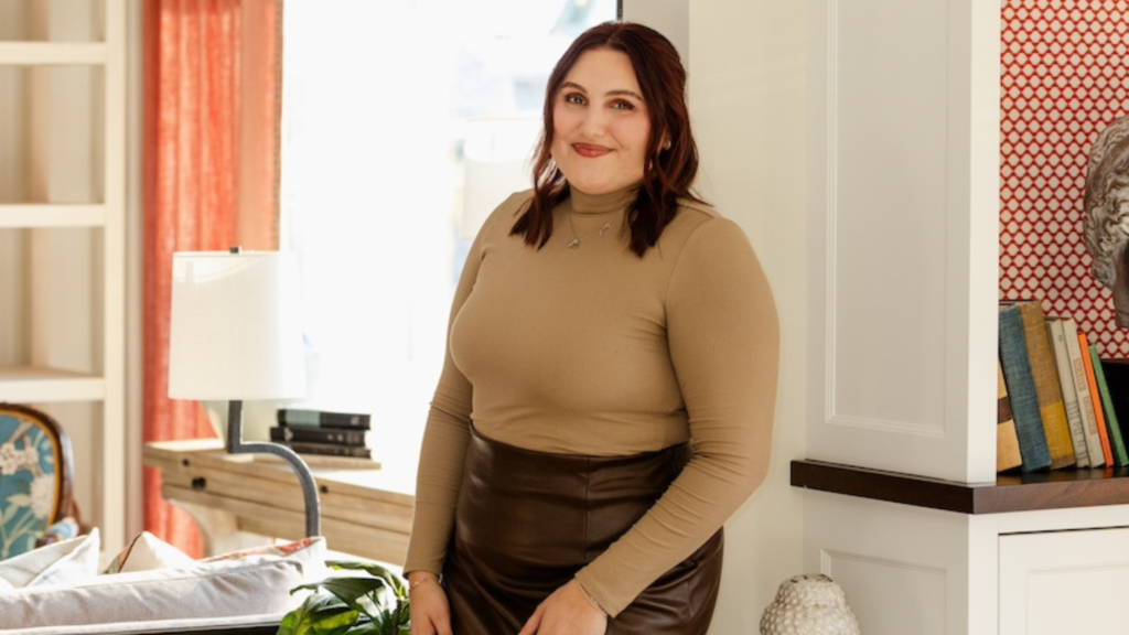 Woman smiling in a warmly lit living room, wearing a tan top and brown skirt. Books and a lamp are in the background. Cozy atmosphere.