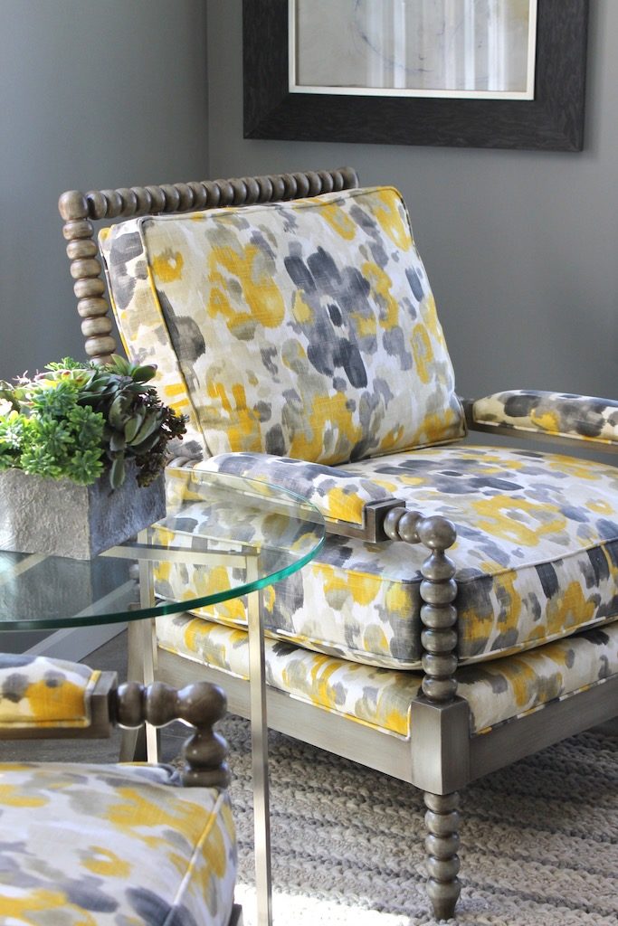 Wooden armchair with a yellow and gray floral cushion beside a round glass table with a potted plant, in a softly lit room.
