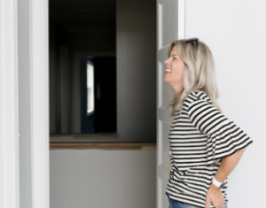 Woman with striped shirt smiling in a bright room, standing near an open doorway. The background is minimal with neutral colors.