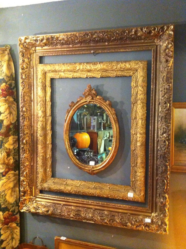 Three nested, ornate, gold picture frames hang on a gray wall. A mirror reflects a pumpkin and room interior. Floral curtain partially visible.