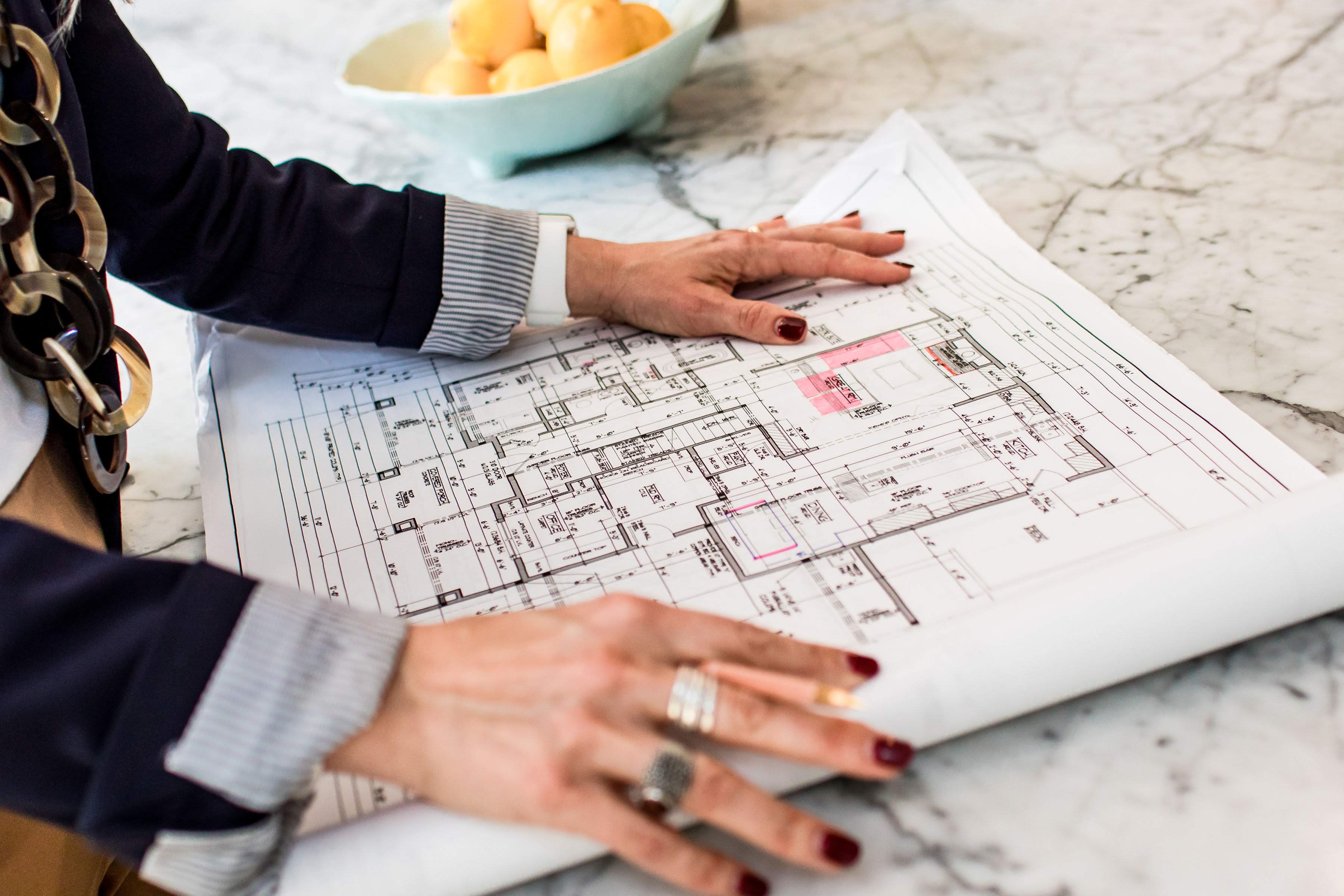 Hands hold architectural plans on a marble table. Bowl of lemons in background. Person wears a navy suit and rings, creating a focused mood.