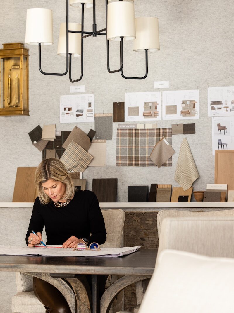 A woman designs at a table with fabric samples and wooden panels on a wall. Neutral colors, modern chandelier, focused atmosphere.