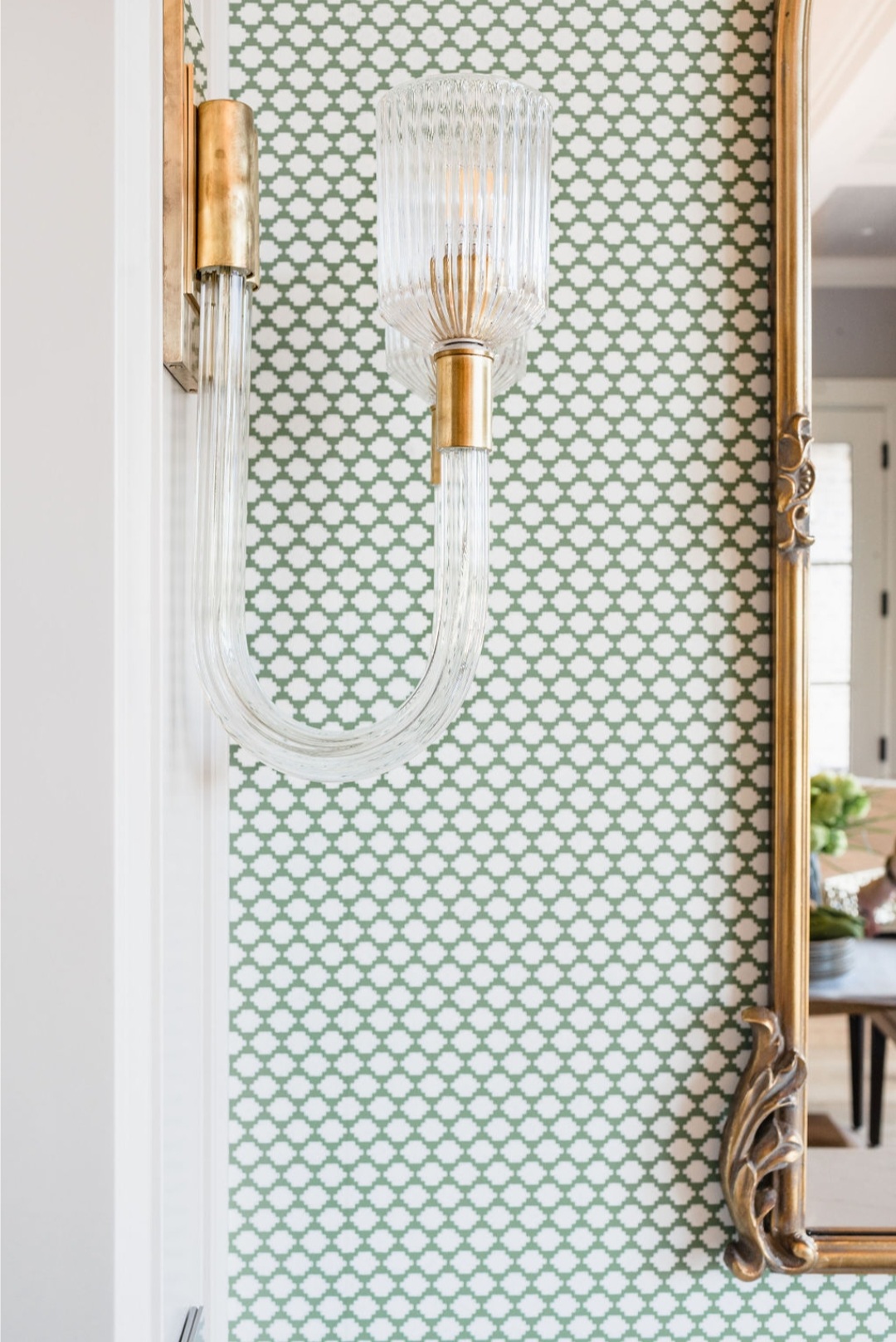 Elegant glass wall sconce with bronze accents on green patterned wallpaper, next to an ornate mirror reflecting a bright room.