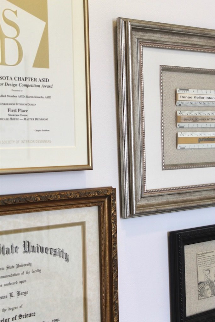 Framed certificates and awards on a white wall. Varied border styles and colors; text details visible. Professional and orderly setting.