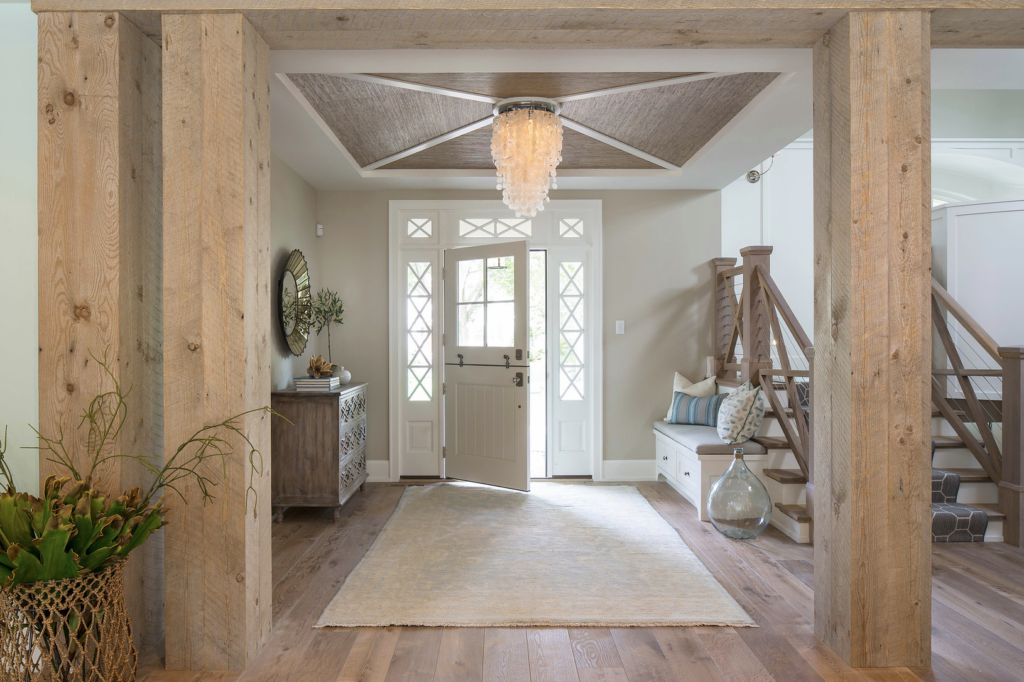 Rustic entryway with wooden beams, a chandelier, open front door, staircase with cushions, and a vase on hardwood floor. Light, airy mood.