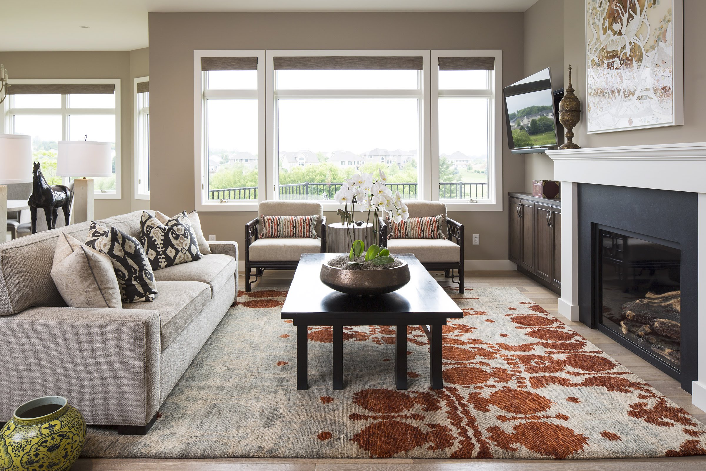Modern living room with a grey sofa, patterned cushions, and a black coffee table with white orchids. Large windows with a scenic view.