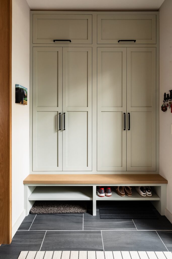 Light green cabinets with black handles in a tidy entryway. Shoes and mats on the tile floor. Calm and organized setting.