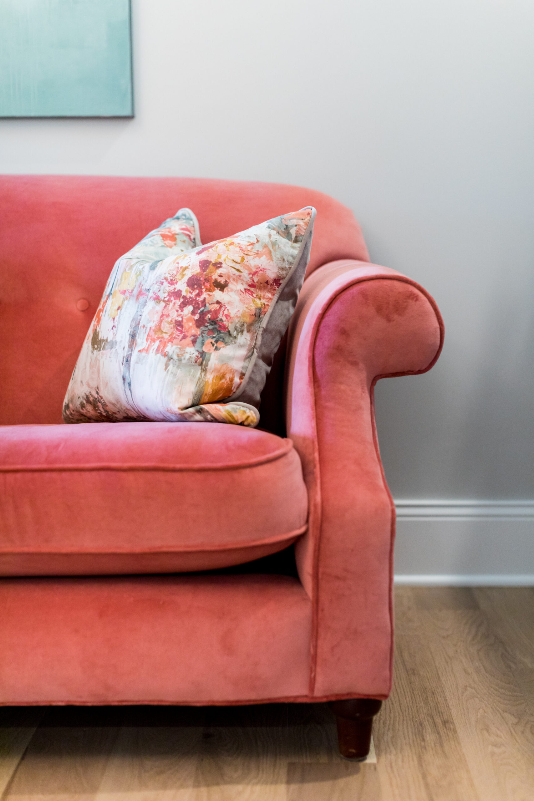 Pink velvet sofa with a colorful floral pillow, set against a light gray wall and wooden floor. A teal artwork is partially visible.