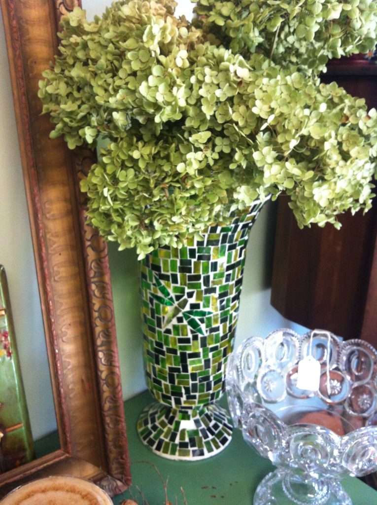 Green hydrangeas in a mosaic vase with tiles of various shades of green, beside an ornate frame and a clear glass bowl with a price tag on a green table.