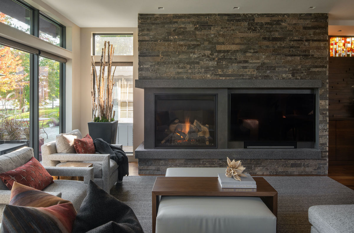 Modern living room with a stone fireplace, beige sofas, and red cushions. Large windows reveal a garden. Warm and cozy ambiance.