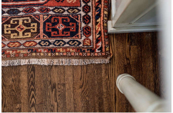 Ornate red and navy patterned rug on dark hardwood floor, adjacent to a white cabinet corner. Rug features geometric designs and fringe.