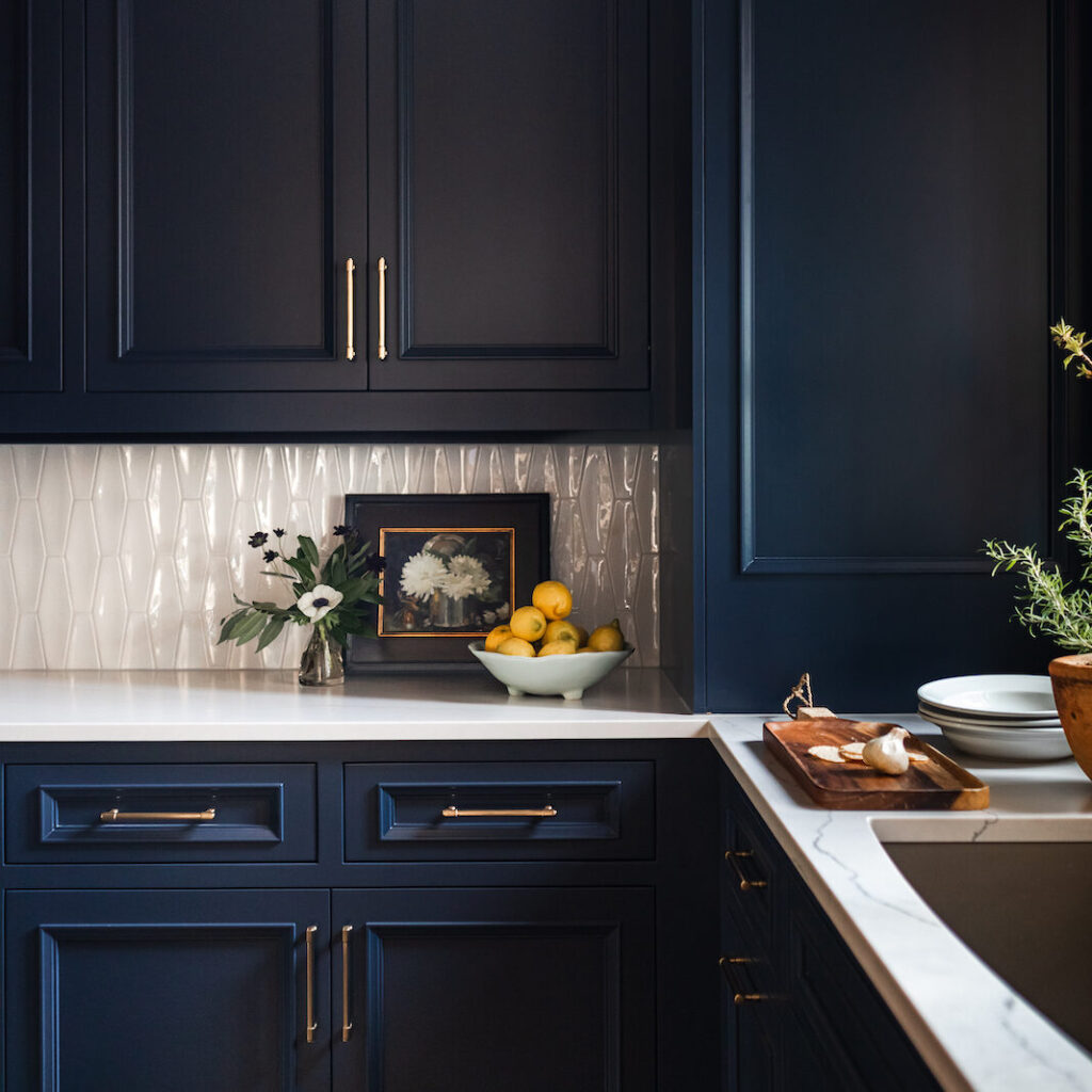 Dark blue kitchen cabinets with gold handles, white countertop, bowl of lemons, small floral vase, and a framed flower picture. Cozy mood.