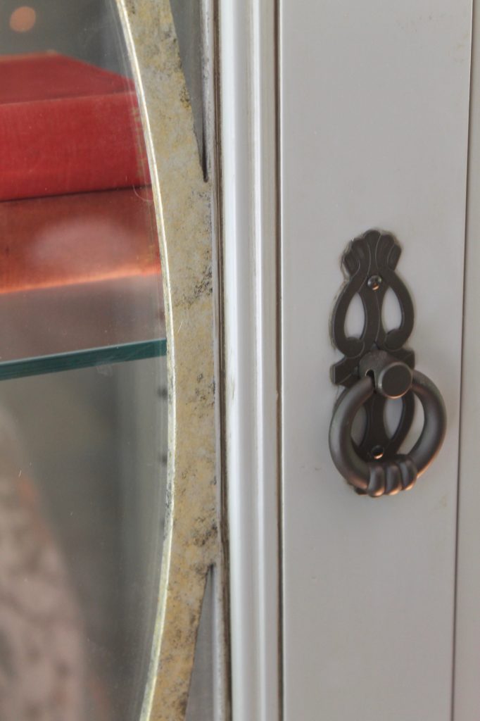 Close-up of a vintage cabinet handle on a pale door with a mirrored section, reflecting a glimpse of red books on a shelf.