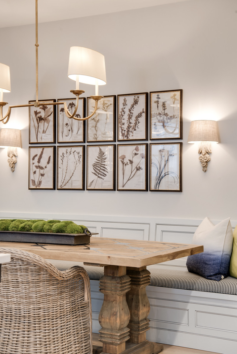 Dining room with a wooden table, wicker chair, and bench with cushions. Wall art of botanical prints and warm lighting create a cozy vibe.