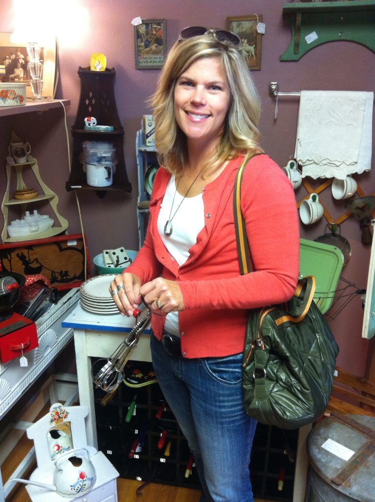 Woman in a vintage shop holding a hand mixer, smiling. Wears an orange cardigan and jeans. Background has dishes, aprons, and framed art. Cozy mood.