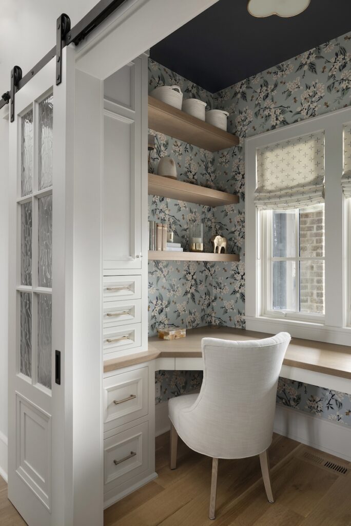 Cozy nook with white chair, floral wallpaper, wooden shelves, pottery, and a window. Light wood floor and sliding glass door enhance charm.