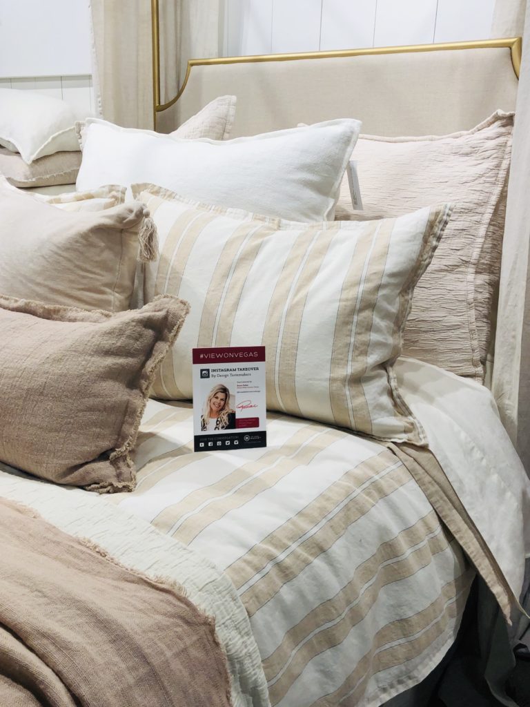 Cozy bed with cream and beige striped pillows and sheets. Sign on bed reads #VIEWONVEGAS and features a woman's face. Relaxing vibe.