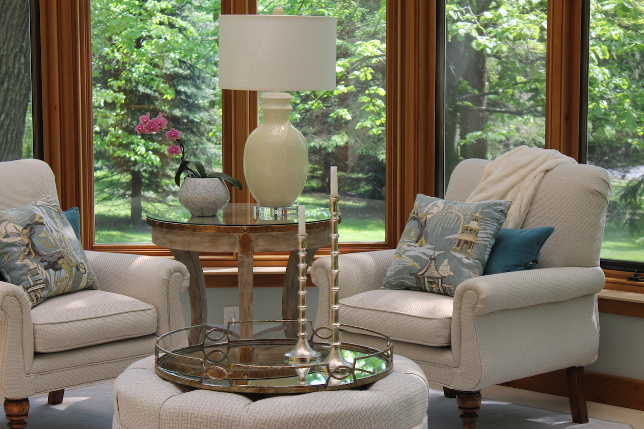 Cozy living room with two beige armchairs, a matching round ottoman with a reflective tray and two candlesticks, a round table with a lamp and a pink orchid. Large windows show lush greenery.