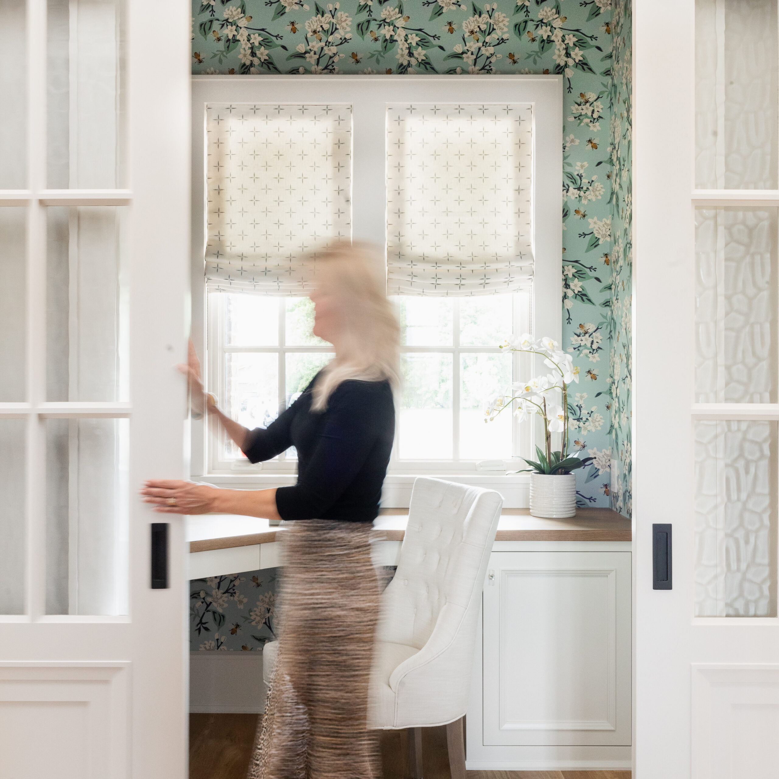 Woman in motion slides door in floral-patterned room with white chair and window. Bright, airy setting with relaxed mood.