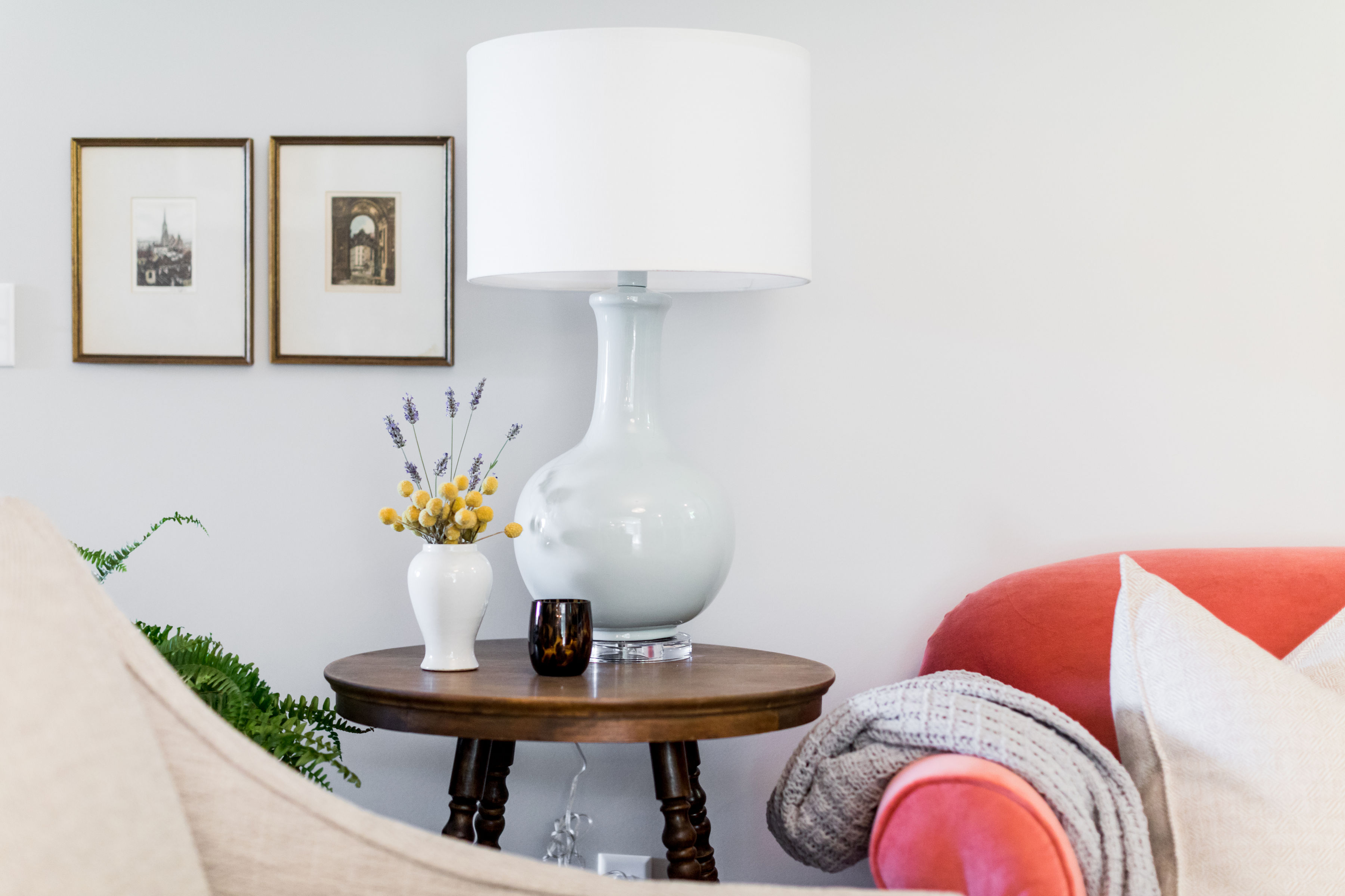 Cozy living room setting with a white lamp, framed art, and flowers on a wooden table. Red couch with a knit blanket adds warmth.