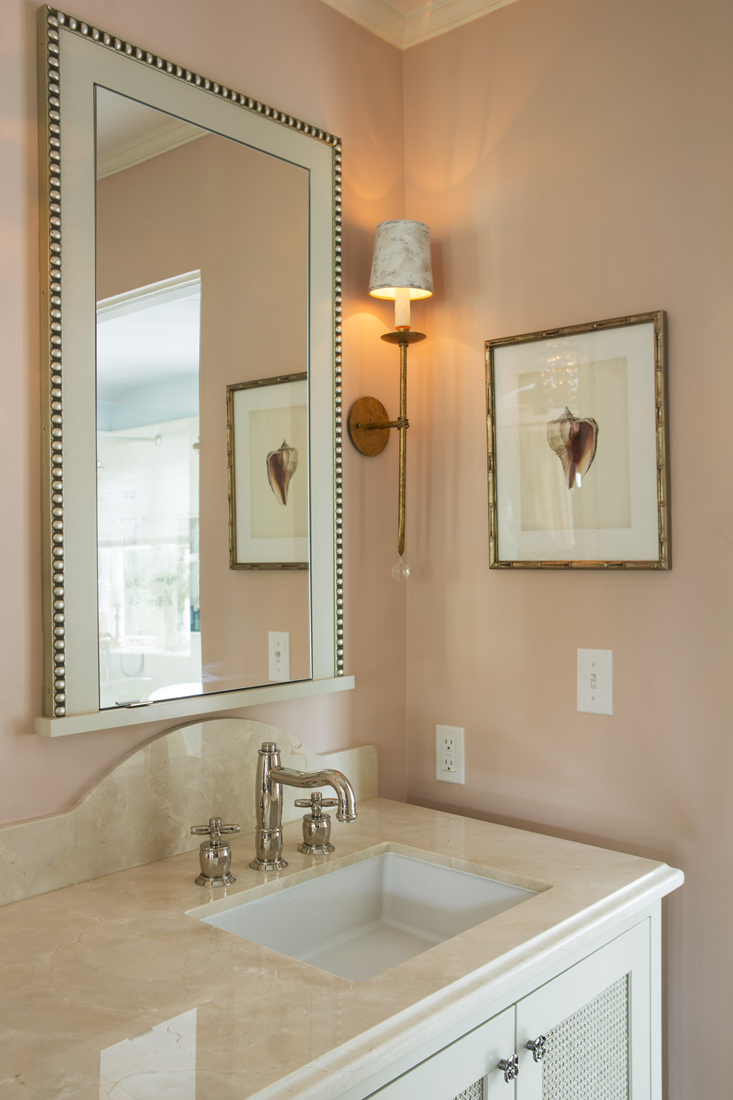 Elegant bathroom with beige walls, large mirror, wall sconce, and shell art. Marble countertop with metal faucet over a white sink.