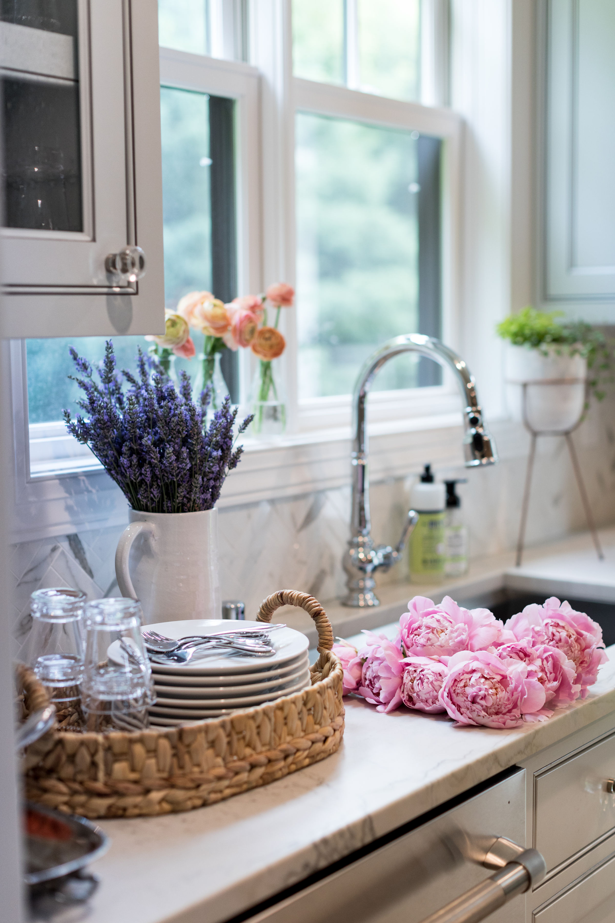 Kitchen with a bright window. Lavender and pink flowers on the counter, dishes in a woven basket, shiny faucet, green soap bottle.