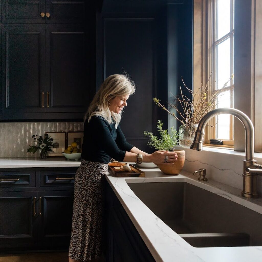 Woman tending to a potted plant by a kitchen sink, sunlight streaming through a window. Dark cabinets, marble countertop, peaceful mood.