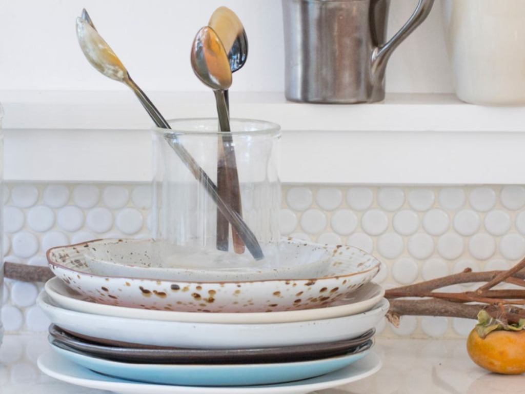 Stacked dishes with a glass jar holding spoons on a white tiled counter. A persimmon and twigs add natural elements. Neutral tones.