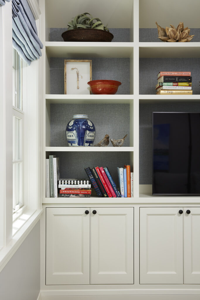White shelves with books, a blue jar, two bird figurines, and a red bowl. A window with blue curtains is on the left. Calm, organized vibe.