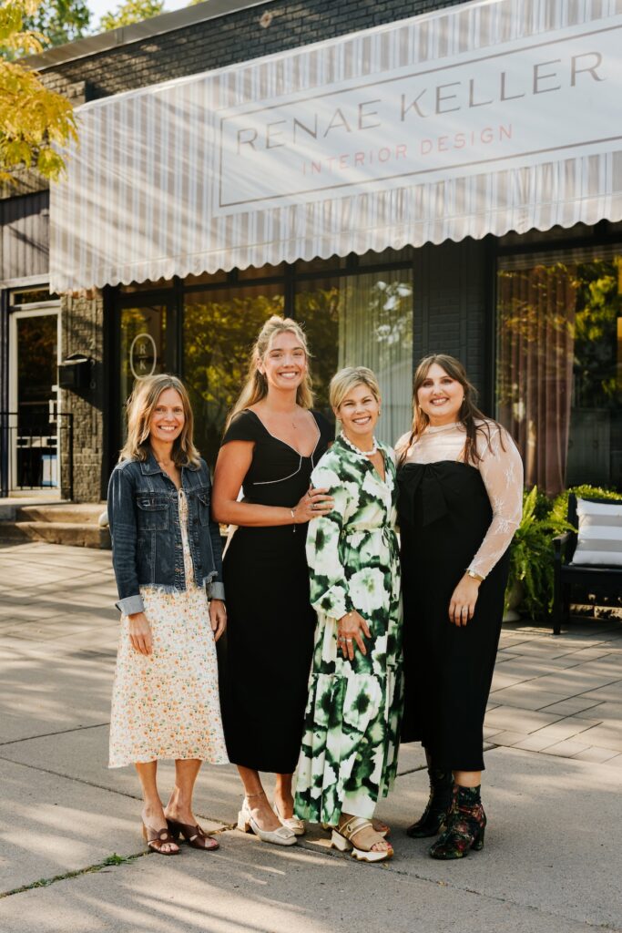 Four women smiling outside "Renae Keller Interior Design." They stand on a sunlit sidewalk, wearing colorful dresses.