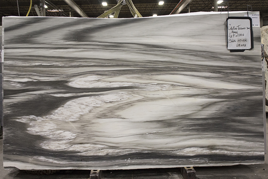 Large marble slab with swirling gray and white patterns in a warehouse. A label with handwritten notes hangs on the upper right corner.