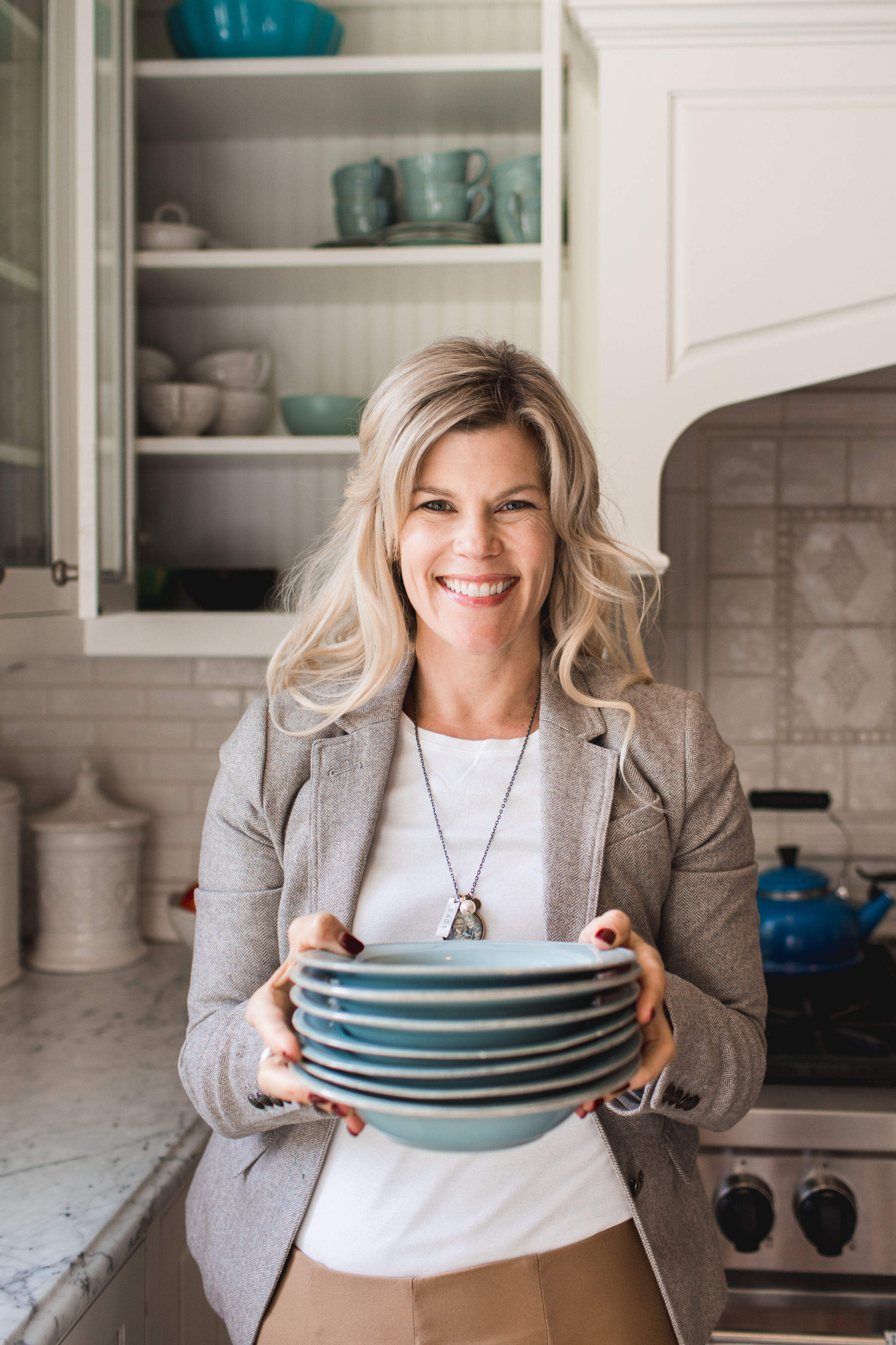 Woman smiling, holding stack of blue bowls in a bright kitchen with white cabinets. Background has teal dishes and marble counters.