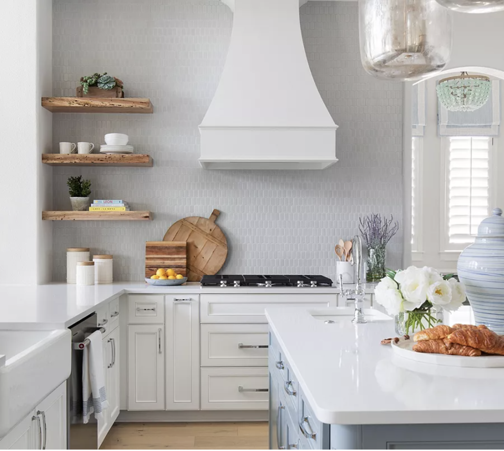 Bright kitchen with white cabinets, wood shelves, and light blue island. Croissants and flowers on counter; lemons, utensils, and decor displayed.