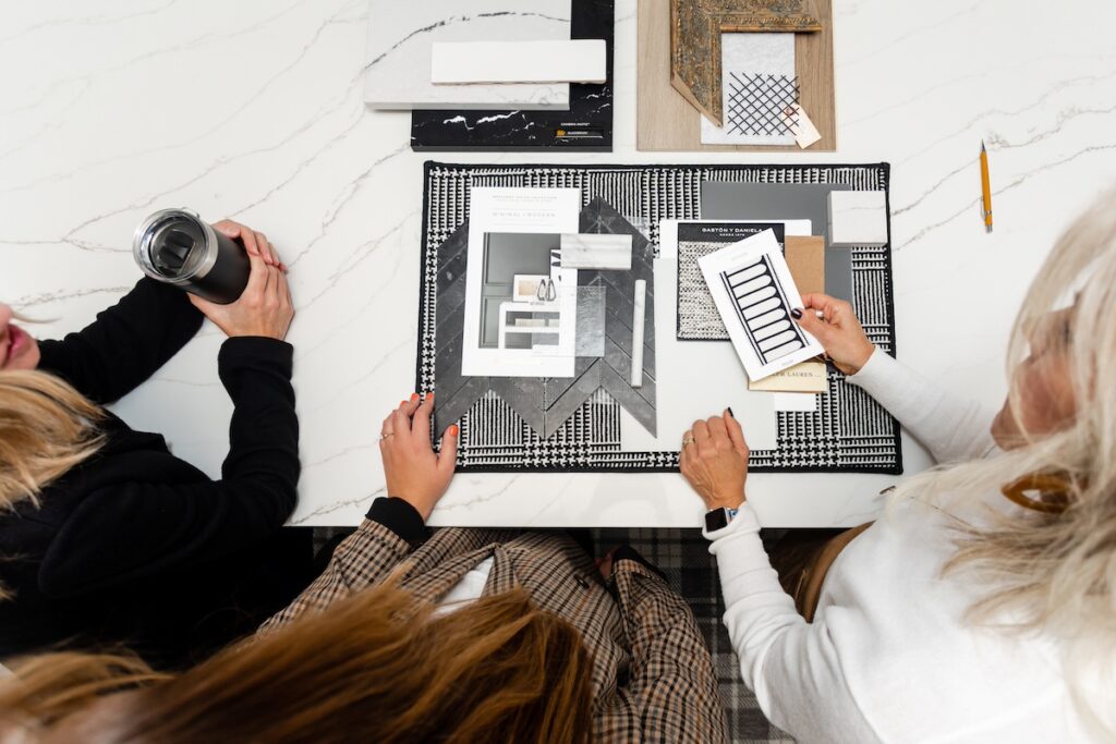 Three people discuss material samples on a patterned table. One holds a black tumbler, another a sample. The setting is bright and organized.