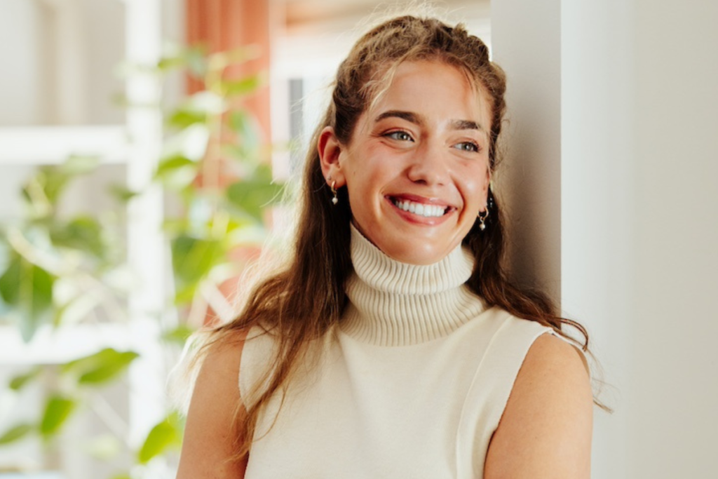 Smiling woman in white turtleneck, leaning against a wall. Green plants and soft pink curtains in the blurred background. Bright, cheerful mood.