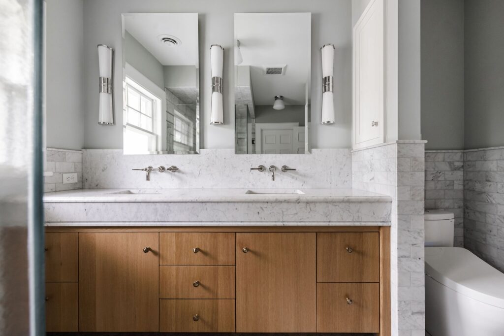 Modern bathroom with dual sinks on marble countertop, wooden cabinets, and two mirrors. Soft natural lighting, gray walls, and minimalist design.