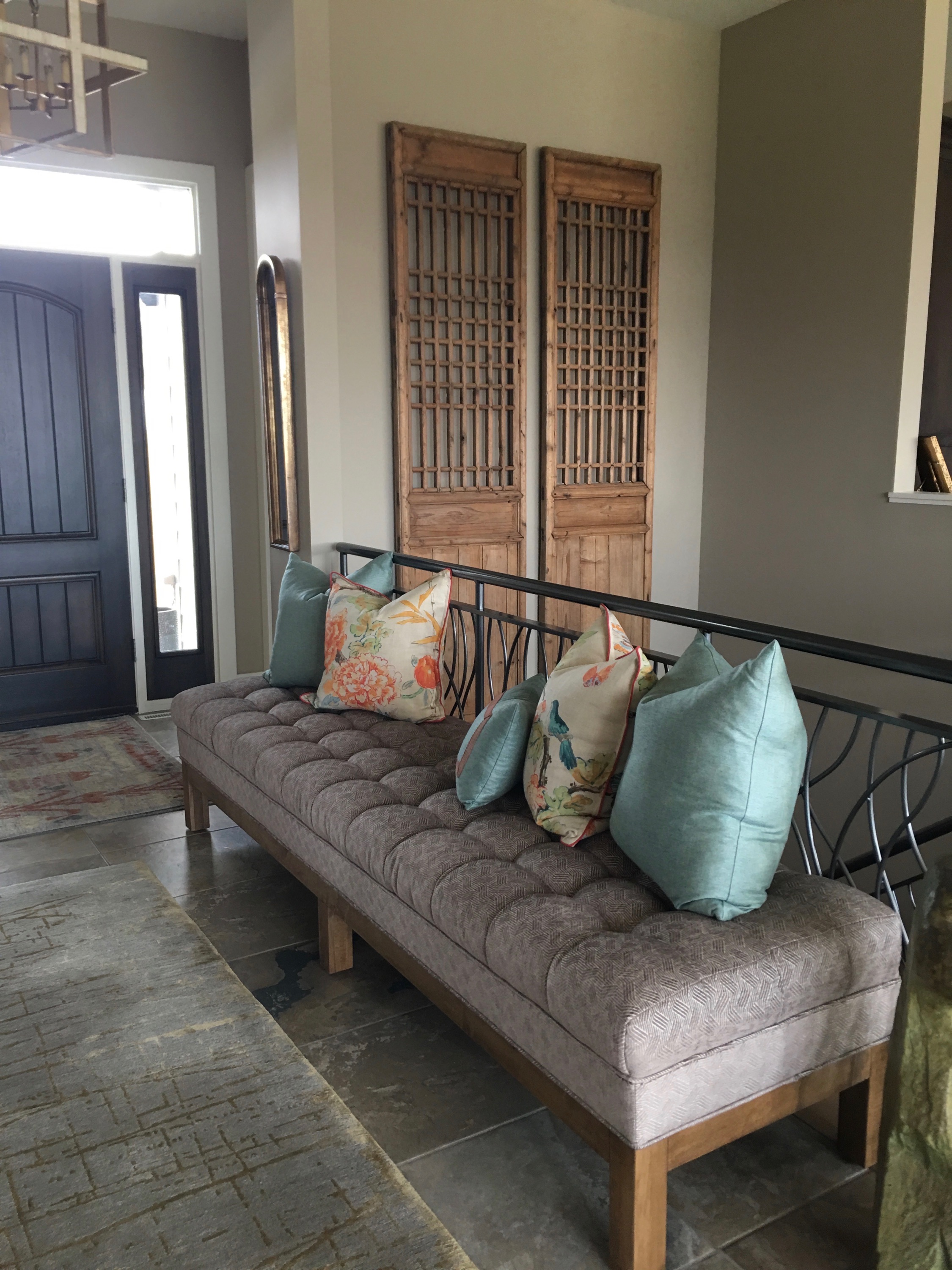 A cozy hallway features a tufted bench with colorful floral and blue pillows, brass wall decor, wooden panels, and a dark front door.