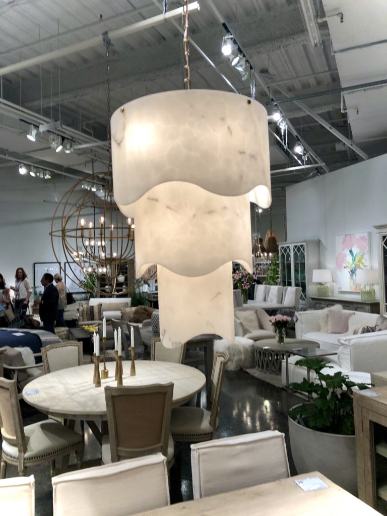 Large white, wavy chandelier in a modern showroom with beige and white furniture. Bright lighting, some people browsing. Calm ambiance.