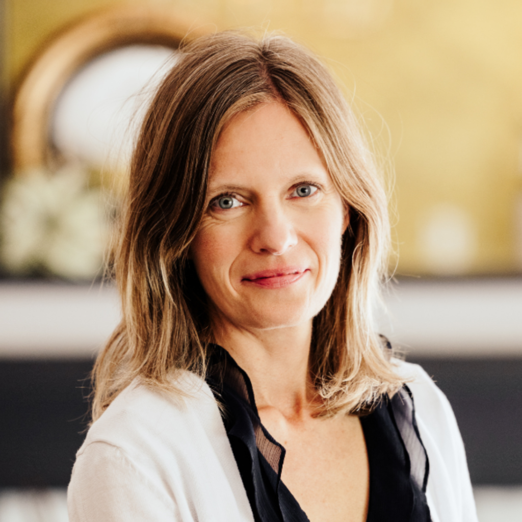 Woman with light brown hair smiles gently. She wears a white top with black trim. Blurred background with warm tones and circular decor.