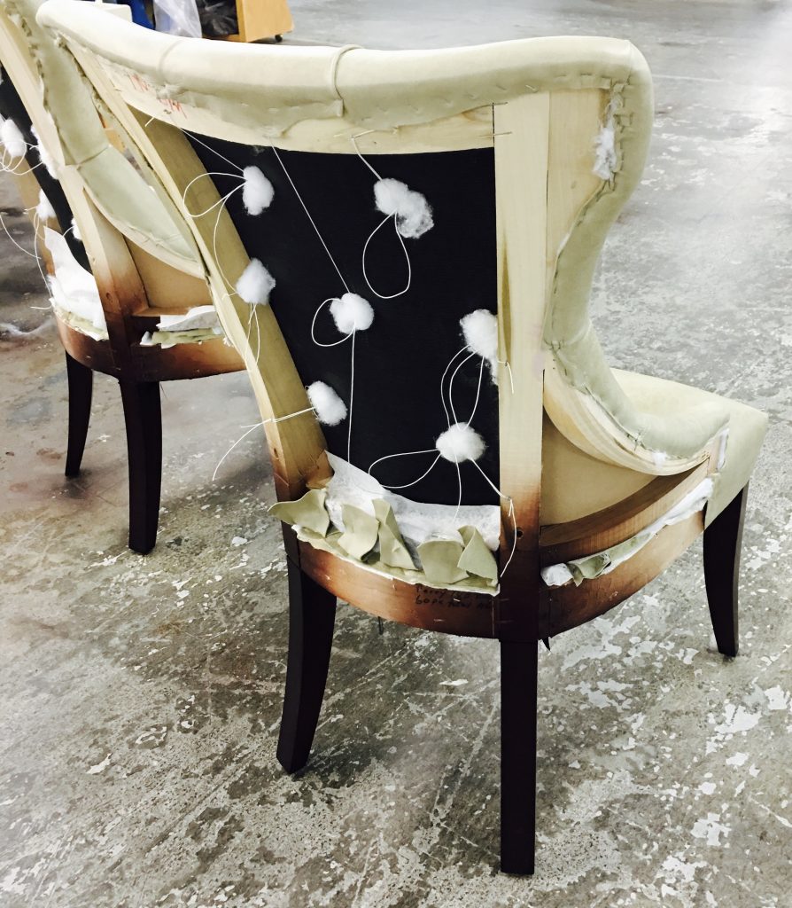 Two unfinished upholstered chairs with exposed stuffing and fabric strips in a workshop. Concrete floor visible, suggesting work in progress.