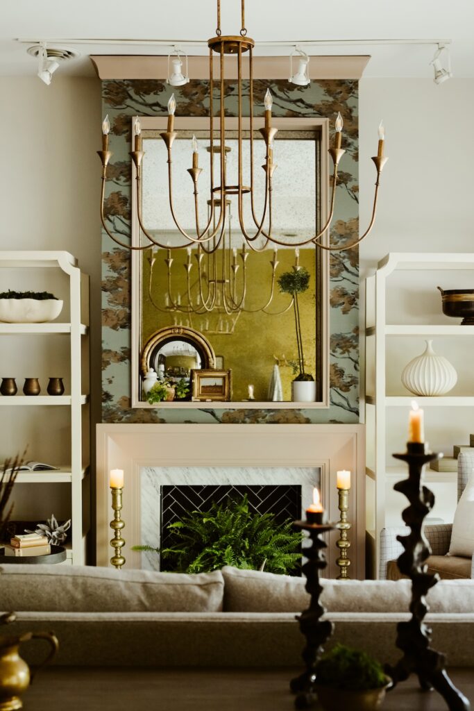 Elegant living room with chandelier, marble fireplace, and green plants. Shelves display vases and candles create a cozy ambiance.