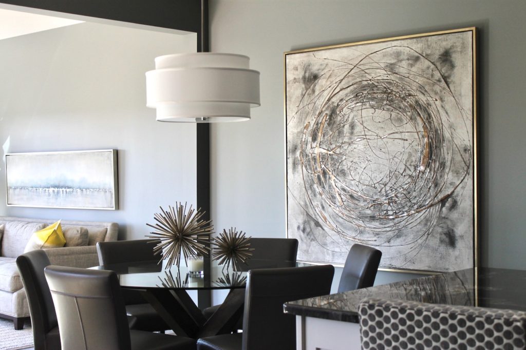 Modern dining room with abstract wall art and spiky metal sculptures on a glass table. Grey chairs, large white pendant light, neutral tones.