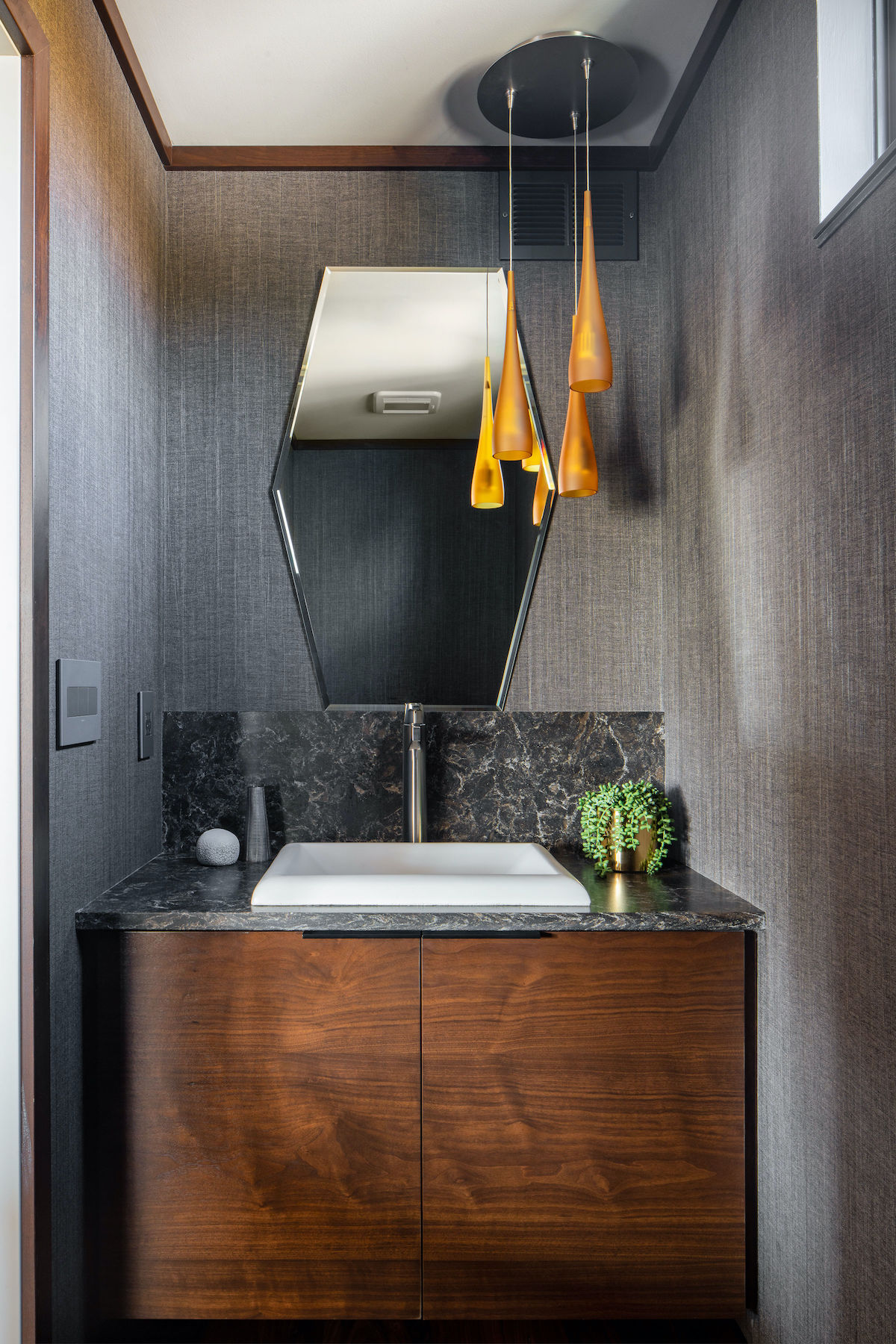 Stylish bathroom with a hexagonal mirror, marble countertop, and wooden vanity. Amber pendant lights and a small potted plant add elegance.