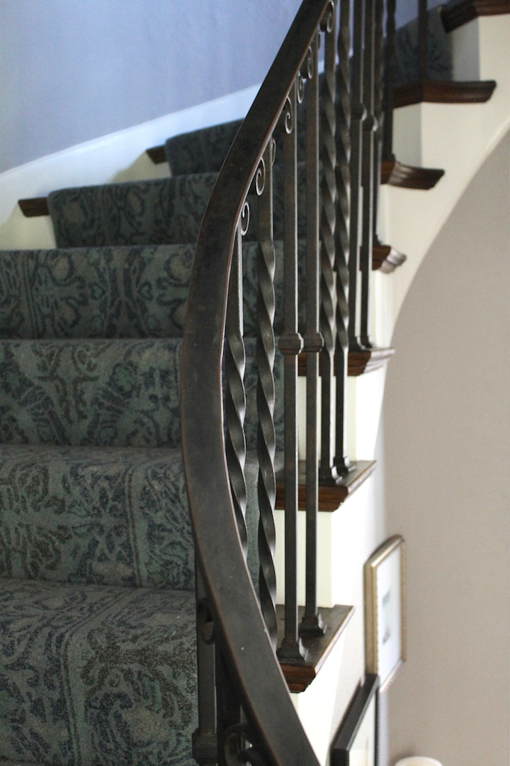 Curved staircase with ornate iron railing. Blue patterned carpet covers steps, framed art on wall. Calm indoor setting.