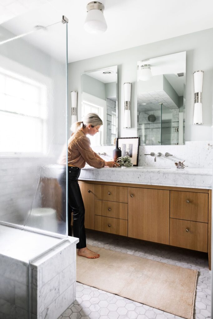 Woman in a bathroom arranging items on a wooden vanity with marble top. Light gray walls, hexagonal tile floor, and large mirrors reflect light.