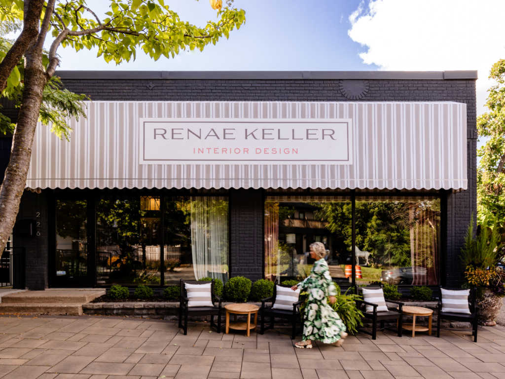 Exterior of "Renae Keller Interior Design" shop with a woman in a floral dress walking past. Black facade, striped awning, patio chairs.