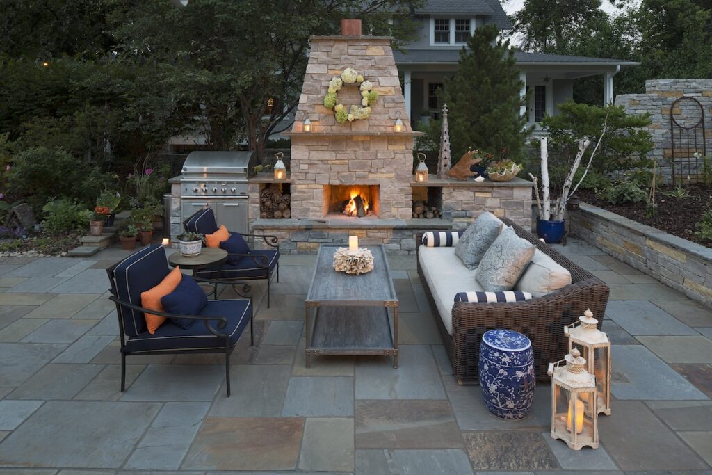 Cozy patio setting with stone fireplace, lit fire, wicker sofa, blue cushions, and lanterns. Trees and house visible in the background.