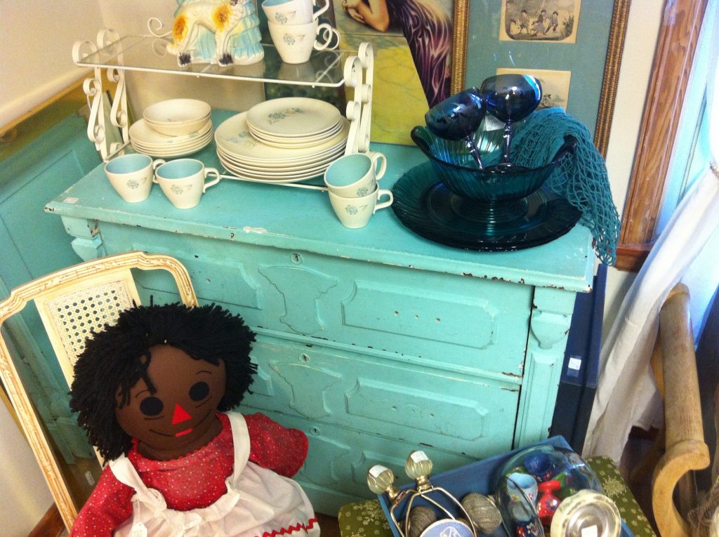Vintage decor with a turquoise cabinet, dishes, glassware, and a Raggedy Ann doll. Warm and nostalgic atmosphere.